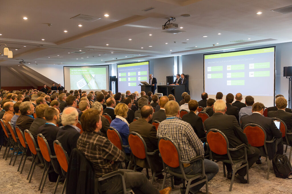 Eine große Gruppe von Personen in Geschäftskleidung sitzt in Reihen vor den Rednern und projizierten ERP-Folien während einer Konferenz oder eines Seminars in einem modernen Konferenzraum.