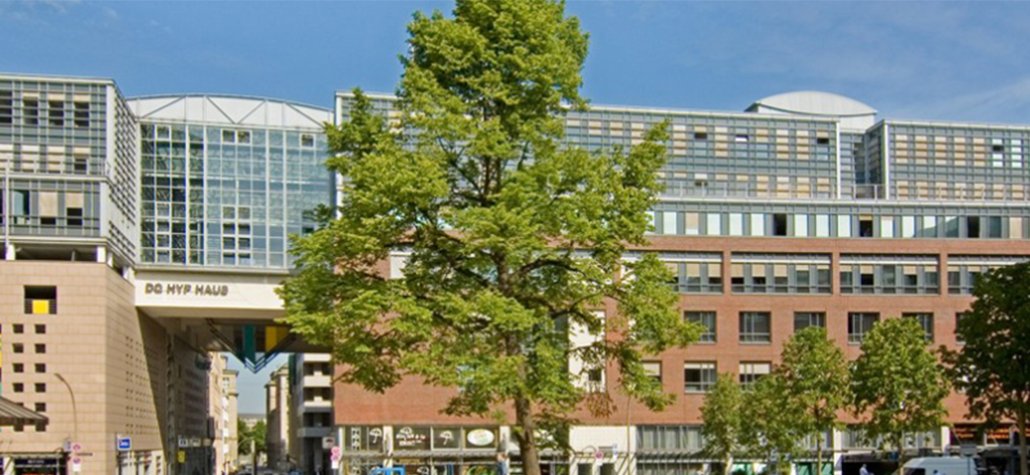 Ein modernes Krankenhausgebäude mit Glas- und Backsteinarchitektur steht an einem sonnigen Tag hinter einem großen, belaubten Baum und mehreren kleineren Bäumen. Der Schriftzug DE UIV MAUD ist auf einem ERP-verbundenen Skywalk zu sehen, der Teile des Gebäudes miteinander verbindet.