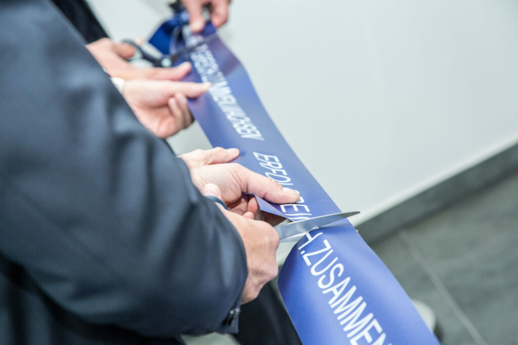 Nahaufnahme von Händen, die ein blaues Band mit einer großen Schere durchschneiden, während einer Zeremonie zum Durchschneiden des Bandes, die die große Eröffnung eines neuen ERP-gestützten Raums oder einer Veranstaltung symbolisiert.