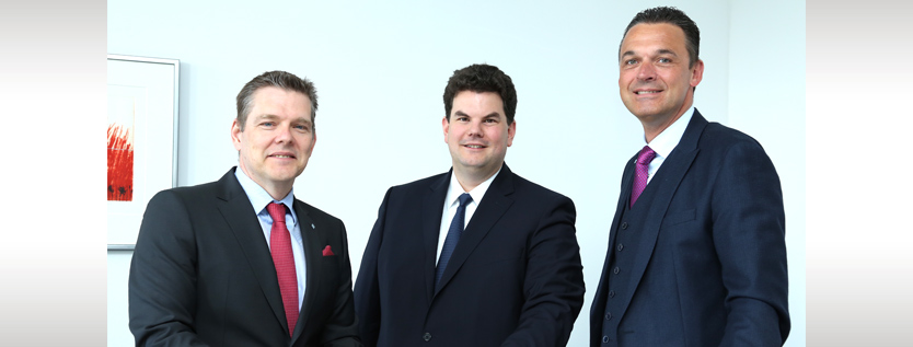 Drei Männer in Geschäftsanzügen stehen zusammen in einem Haus und lächeln in die Kamera. Der Mann auf der rechten Seite schüttelt dem Mann in der Mitte die Hand und feiert eine erfolgreiche ERP-Partnerschaft. Ein gerahmtes Kunstwerk hängt an der weißen Wand hinter ihnen.