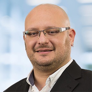 Ein kahlköpfiger Mann mit Brille und kurzem Bart, der ein weißes Hemd und einen dunklen Blazer trägt, lächelt vor einem unscharfen blau-weißen Hintergrund in die Kamera und strahlt das Selbstbewusstsein eines ERP-Profis aus.