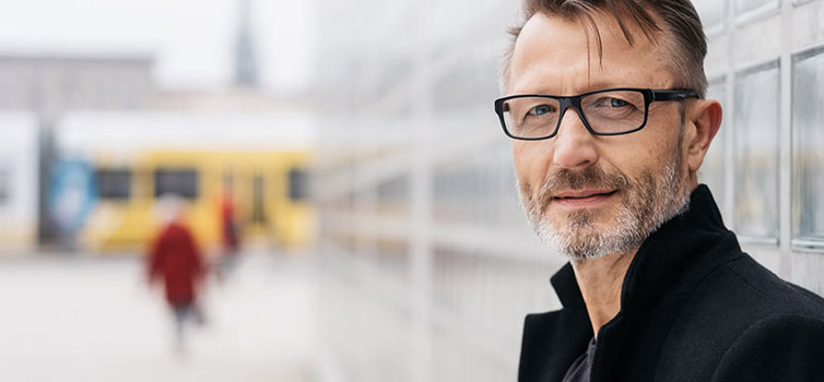 Ein Mann mittleren Alters mit kurzen Haaren, Bart und Brille steht draußen vor einer Glaswand und trägt einen schwarzen Mantel. Im Hintergrund sind eine verschwommene gelbe Straßenbahn und Menschen zu sehen, was auf einen ERP-Berater schließen lässt, der in einer städtischen Umgebung unterwegs ist.