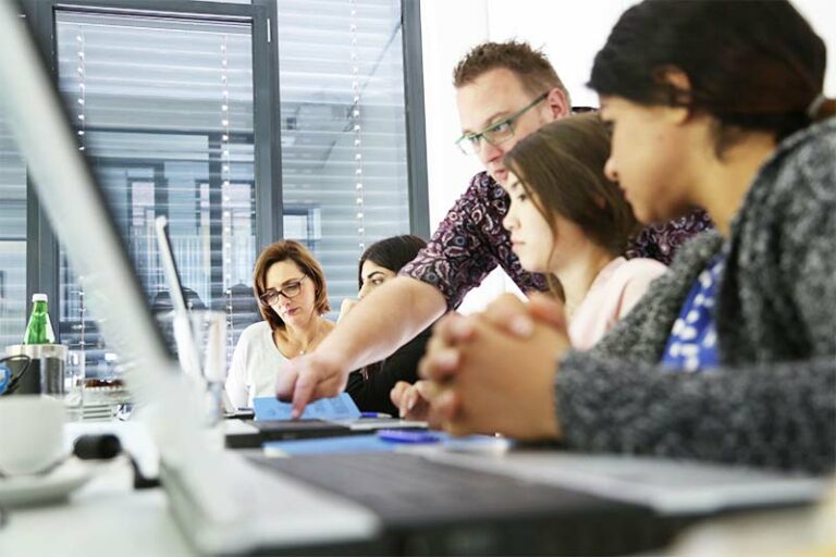 Eine Gruppe von Personen sitzt um einen Konferenztisch herum, konzentriert auf ERP-Dokumente und Laptops, während eine Person aufsteht und während einer gemeinsamen Sitzung in einem hellen Büro auf ein Papier zeigt.
