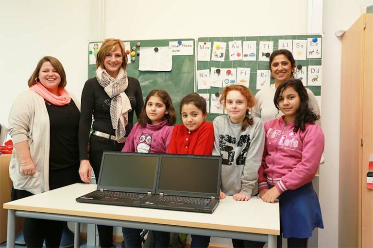 Vier junge Mädchen stehen vor einem Tisch mit zwei geöffneten Laptops, auf denen ein ERP-System abgebildet ist, begleitet von drei erwachsenen Frauen. Alle lächeln. Hinter ihnen steht eine grüne Kreidetafel mit bunten Zeichnungen und Buchstaben.