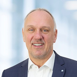 Ein lächelnder Mann mittleren Alters mit kurzen hellen Haaren und gestutztem Bart, der ein weißes Hemd und eine dunkle Anzugsjacke trägt, steht vor einem unscharfen hellblauen Hintergrund - perfekt für die Darstellung von Fachwissen über ERP-Lösungen.