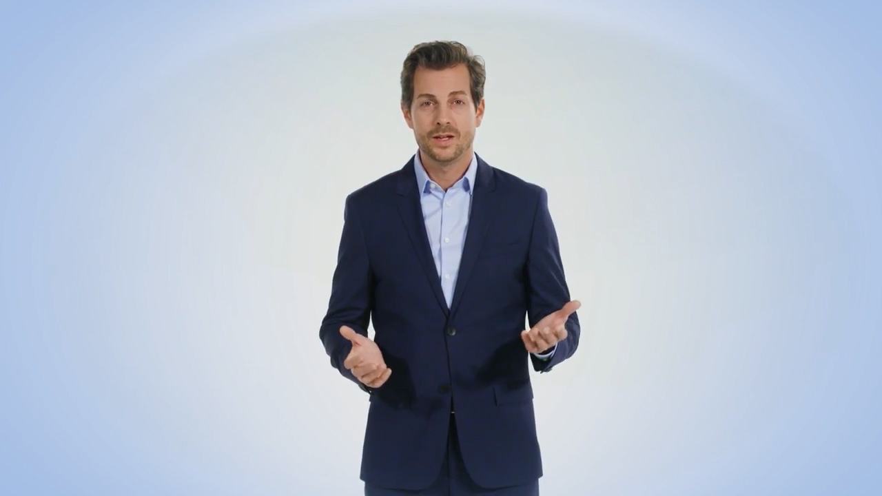 Ein Mann in einem marineblauen Anzug und einem hellblauen Hemd steht vor einem einfarbigen, hellen Hintergrund und gestikuliert mit beiden Händen, als würde er eine ERP-Lösung präsentieren.