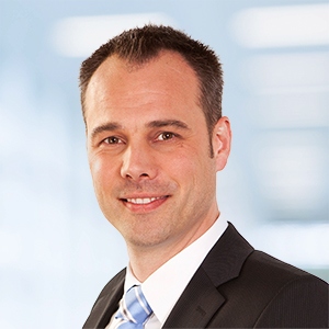 Ein Mann in dunklem Anzug, weißem Hemd und blau gestreifter Krawatte lächelt vor einem unscharfen hellblauen Hintergrund in die Kamera und strahlt Selbstbewusstsein und Professionalität aus, wie man sie häufig bei ERP-Spezialisten findet.