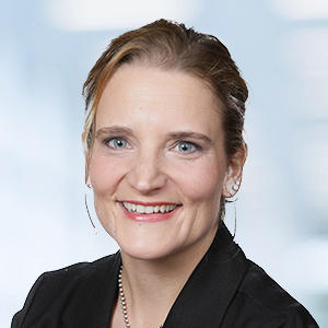 Eine Frau mit heller Haut und blauen Augen lächelt in die Kamera. Sie hat ihr hellbraunes Haar zurückgesteckt und trägt einen schwarzen Blazer, eine Halskette und kleine Ohrringe - genau richtig für ihre Rolle als ERP-Spezialistin. Der Hintergrund ist leicht unscharf und in hellen Farben gehalten.