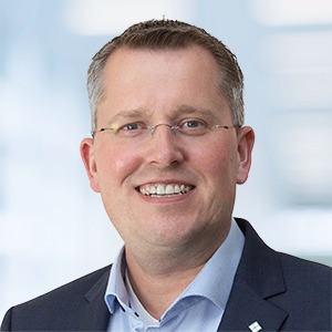 Ein lächelnder Mann mit kurzen hellbraunen Haaren und Brille, der einen marineblauen Blazer über einem hellblauen Hemd mit Kragen trägt, steht vor einem unscharfen hellen Hintergrund und strahlt Selbstbewusstsein als Experte für ERP-Lösungen aus.