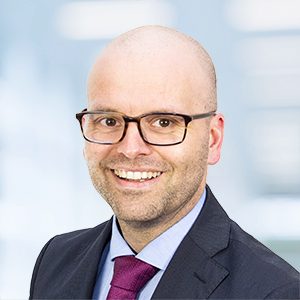 Ein lächelnder Mann mit Brille und Glatze, gekleidet in einen dunklen Anzug, ein hellblaues Hemd und eine weinrote Krawatte, posiert vor einem unscharfen, hellen Hintergrund.