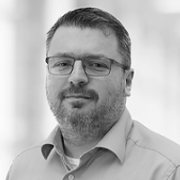 Ein Mann mit kurzen Haaren, Brille und Bart trägt ein Hemd mit Kragen und schaut mit einem leichten Lächeln in die Kamera. Der unscharfe, helle Hintergrund verleiht dem Porträt die Ausstrahlung eines professionellen ERP-Beraters.