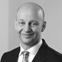 Ein glatzköpfiger Mann in Anzug und Krawatte lächelt in die Kamera vor einem schlichten Hintergrund. Das Schwarz-Weiß-Foto hat einen professionellen Ton, perfekt für Geschäftsprofile oder ERP-Führungsporträts.