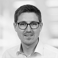 Ein Mann mit kurzen Haaren und Brille, der ein helles Hemd mit Kragen trägt, lächelt leicht in einem Schwarz-Weiß-Porträt mit unscharfem Hintergrund, das die Zuversicht eines ERP-Experten widerspiegelt.