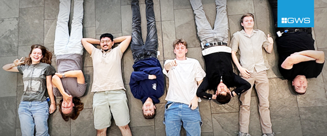 GWS-Lehrlinge2025 Sieben junge Erwachsene liegen nebeneinander auf einem Steinboden, lächeln und machen spielerische Gesten. In der oberen rechten Ecke erscheint ein blaues rechteckiges Logo mit GWS.