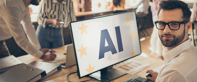 KI-Schulungen-2 Ein Mann mit Brille sitzt an einem Schreibtisch und blickt in die Kamera. Auf einem Computermonitor sind AI und gelbe Sterne zu sehen. Im Hintergrund arbeiten Menschen an einem Tisch in einer modernen Büroumgebung zusammen.
