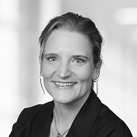 Schwarz-Weiß-Porträt einer lächelnden Frau mit hellen, zurückgesteckten Haaren, die einen Blazer und Reifohrringe trägt, vor einem unscharfen Hintergrund.