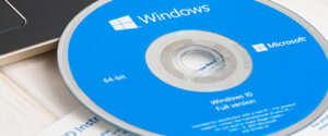 Ein blauer Windows 10-Installationsdatenträger mit den Aufschriften "64-Bit" und "Vollversion" liegt teilweise im CD-Laufwerk eines Laptops, darunter ist ein Teil eines Papierdokuments sichtbar.