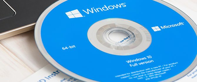 Ein blauer Windows 10-Installationsdatenträger mit den Aufschriften "64-Bit" und "Vollversion" liegt teilweise im CD-Laufwerk eines Laptops, darunter ist ein Teil eines Papierdokuments sichtbar.