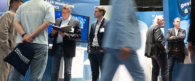 Menschen interagieren und gehen durch einen belebten Messe- oder Konferenzstand mit blauen Displays und Branding im Hintergrund. Einige stehen, sprechen oder lesen, während andere leicht verschwommen vorbeilaufen.