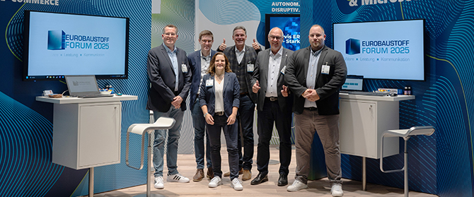 Sechs Personen stehen lächelnd in einem Messestand mit Eurobaustoff Forum 2025-Schildern, tragen Geschäftskleidung und Konferenzabzeichen, mit blau-weißem Display-Hintergrund und Barhockern daneben.