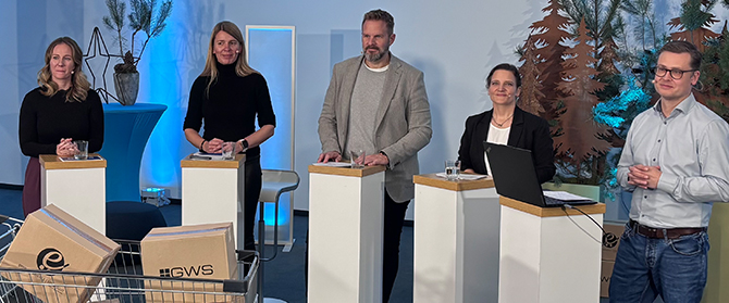 Fünf Personen stehen hinter Podien in einer Studiokulisse, mit dekorativen Bäumen und blauer Beleuchtung im Hintergrund. Im Vordergrund steht ein Einkaufswagen mit Pappkartons, und auf einem Podest steht ein Laptop.