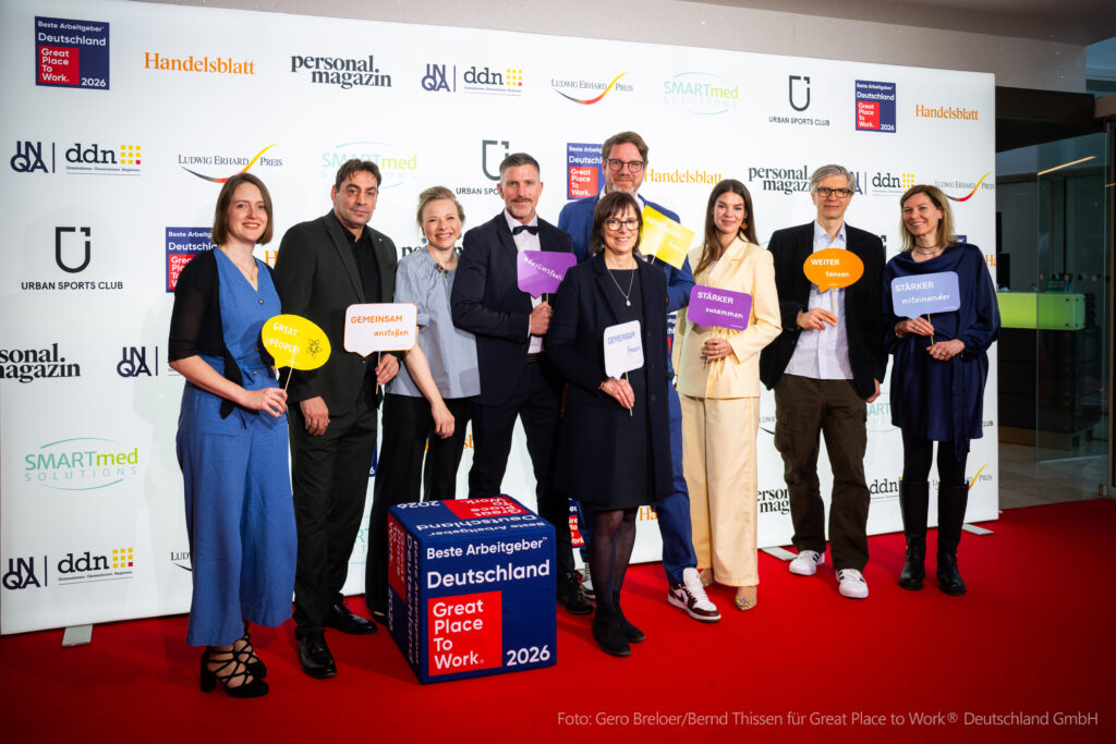 Neun Personen stehen auf einem roten Teppich mit bunten Sprechblasenschildern und posieren für ein Gruppenfoto bei einer Veranstaltung von "Great Place to Work Deutschland 2026". Im Hintergrund sind Markenbanner und -logos zu sehen.