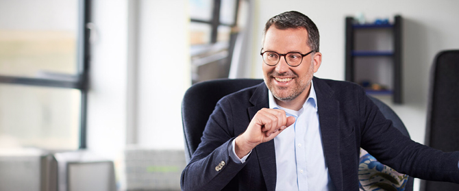 Ein Mann mit Brille und Bart, der einen dunklen Blazer über einem hellen Hemd trägt, lächelt und gestikuliert mit seiner rechten Hand, während er in einem modernen, hell erleuchteten Büroraum sitzt.