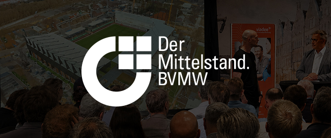Ein großes weißes Logo und der Schriftzug „Der Mittelstand. BVMW“ überlagern ein Publikum, das zwei Rednern auf der Bühne zusieht, während im Hintergrund schemenhaft ein Stadion und ein Stadtgebiet zu erkennen sind.