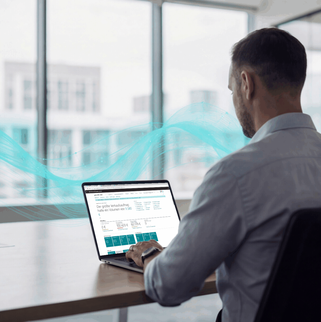 Ein Mann in einem hellblauen Hemd sitzt an einem Schreibtisch und arbeitet an einem Laptop in einem hellen Büro mit großen Fenstern. Darüber sind blaue digitale Wellen zu sehen, die auf Technologie oder Vernetzung hindeuten.