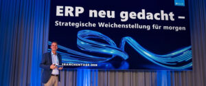 Ein Mann steht auf der Bühne und hält ein Tablet vor eine große Leinwand, auf der der deutsche Text „ERP neu gedacht – Strategische Weichenstellung für morgen“ zu sehen ist, umrahmt von blauen digitalen Grafiken und Vorhängen im Hintergrund.