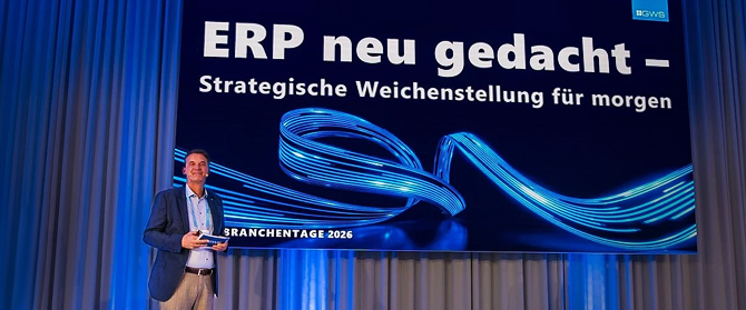Ein Mann steht auf der Bühne und hält ein Tablet vor eine große Leinwand, auf der der deutsche Text „ERP neu gedacht – Strategische Weichenstellung für morgen“ zu sehen ist, umrahmt von blauen digitalen Grafiken und Vorhängen im Hintergrund.