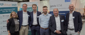 Fünf Personen stehen in einer Reihe an einem Messestand vor Firmenbannern.